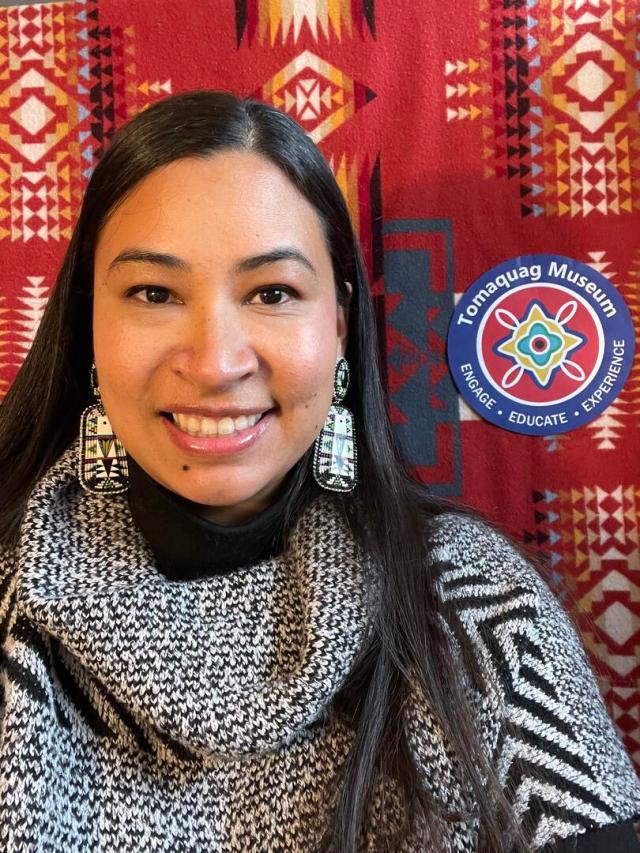 Closeup image of speaker and storyteller Silvermoon Mars LaRose. She has long, dark hair and wears large, black-and-white earrings with intricate patterns. She wears a black and white shawl. She stands in front of a red background with triangular patterns, and a sticker that says "Tomaquag Museum: Engage, Educate, Experience" sits on the background to Silvermoon's right.